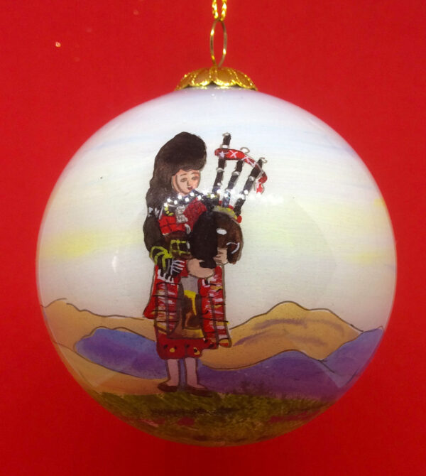 A beautiful handpainted bauble, decorated with an images of a Scottish lone piper in full Highland regalia. He stands upon a Scottish hillside with mountains in the background.