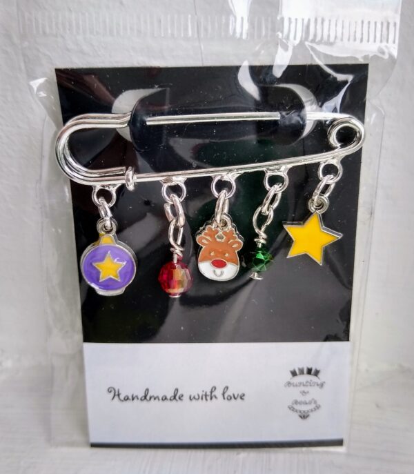 A Festive Brooch, made from a silver kiltpin with festive charms including a purple bauble with yellow star, a red faceted beaad, a cute reindeer face, a green faceted bead and a yellow star. Hand made with love by Bunting & Beads.