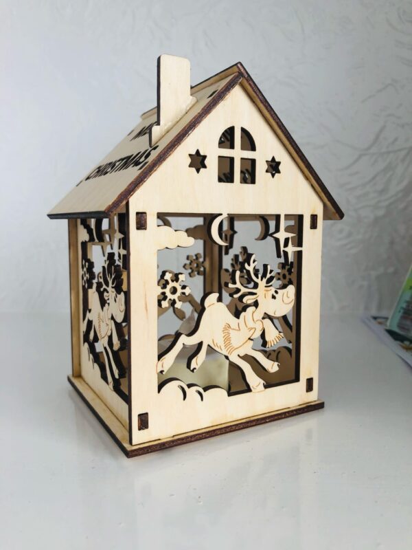 A 3D laser cut wooden tea light holder in the shape of a house. The words 'Merry Christmas' have been cut out of the roof and a cut out leaping reindeer design can be seen on the sides of the house.