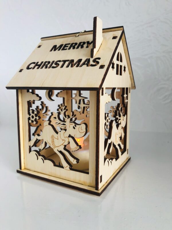 A 3D laser cut wooden tea light holder in the shape of a house. The words 'Merry Christmas' have been cut out of the roof and a cut out leaping reindeer design can be seen on the sides of the house. A battery operated tea light illuminates from with in.