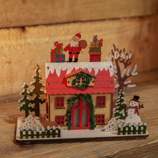 An unlit LED Wooden Christmas Cottage with a Santa Claus on top. This cute wooden cottage had a lovelty green garland around it's red door which is flanked by four square red windows. The dormer window in the red roof is decorated with a matching green wreath. On top of the red roof tile is a covering os snow decorated with iridescent glitter upon which Sanat Claus stands with his gift bag and two stacks of presents. The cottage sits in a garden with snowy trees and shrubs and a cute picket fence. In the garden there is a small brown deer and a cheerful snowman.