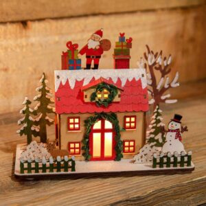 A LED Wooden Christmas Cottage with a Santa Claus on top. This cute wooden cottage had a lovelty green garland around it's red door which is flanked by four square red windows. The dormer window in the red roof is decorated with a matching green wreath. Warm white light is shining out from the windows and door. On top of the red roof tile is a covering os snow decorated with iridescent glitter upon which Sanat Claus stands with his gift bag and two stacks of presents. The cottage sits in a garden with snowy trees and shrubs and a cute picket fence. In the garden there is a small brown deer and a cheerful snowman.