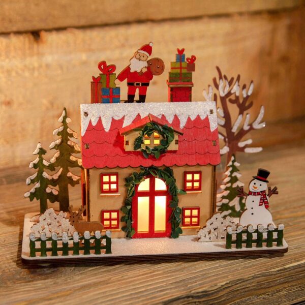 A LED Wooden Christmas Cottage with a Santa Claus on top. This cute wooden cottage had a lovelty green garland around it's red door which is flanked by four square red windows. The dormer window in the red roof is decorated with a matching green wreath. Warm white light is shining out from the windows and door. On top of the red roof tile is a covering os snow decorated with iridescent glitter upon which Sanat Claus stands with his gift bag and two stacks of presents. The cottage sits in a garden with snowy trees and shrubs and a cute picket fence. In the garden there is a small brown deer and a cheerful snowman.