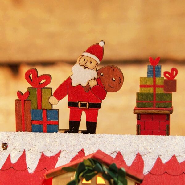 A close up of Santa from our LED Wooden Christmas Cottage with a Santa Claus on top. This cute wooden cottage had a lovelty green garland around it's red door which is flanked by four square red windows. The dormer window in the red roof is decorated with a matching green wreath. On top of the red roof tile is a covering os snow decorated with iridescent glitter upon which Sanat Claus stands with his gift bag and two stacks of presents. The cottage sits in a garden with snowy trees and shrubs and a cute picket fence. In the garden there is a small brown deer and a cheerful snowman.