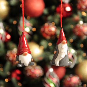 Two Ceramic Hanging Nordic Gonks hanging from red ribbons. Both have red hats and grey bodies, pink noses and hands and white beards. The one on the left is short and dumpy while the one on the right tall and elongated.