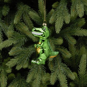 A traditional blown glass Christmas tree decoration in the shape of a cute T-Rex dinosaur. This dinosaur is decorated in bright green metallic paint with yellow metallic paint on his tummy and under his tail. His teeth are bright white and he has green glitter decoration running down his tail from the top of his head. He hands on a gold string.