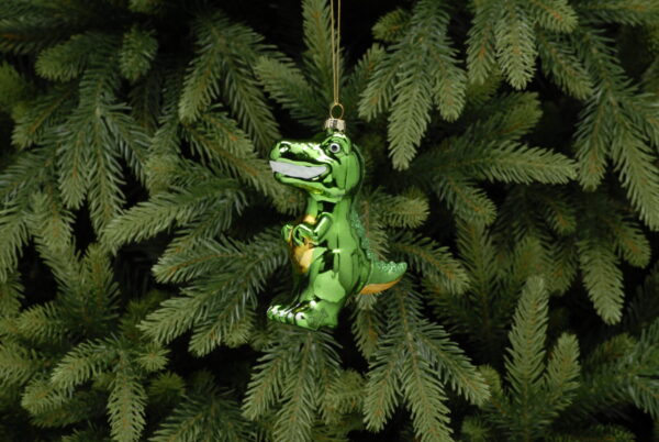 A traditional blown glass Christmas tree decoration in the shape of a cute T-Rex dinosaur. This dinosaur is decorated in bright green metallic paint with yellow metallic paint on his tummy and under his tail. His teeth are bright white and he has green glitter decoration running down his tail from the top of his head. He hands on a gold string.