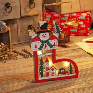 An LED Wooden Ornament in the shape of a boot. The inside of this wooden boot has a die cut painted wooden festive scene depicting a white church and some green fir trees next to a red car laden with a Christmas tree beneath a cresecent moon and clouds. Behind this scene is an opaque plastic layer behind which sits the warm white LED lights that glow through the die cut scene. The out side of the wooden boot is red and the cuff of the boot is white painted with red stitching deatil and three red Christmas tree shapes. A snowman is poking out of the cuff with his arms in the air beside his head. He is wearinga green scarf and black top hat. He has a cheerful expression and an orange carrot nose. He is holding a green Christmas tree in his left hand.