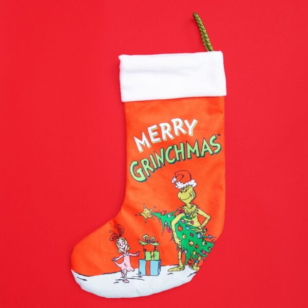 A red fabric Christmas stocking with a white cuff and gold cord for hanging. The body of the stocking features an illustration of the Grinch stealing a Christmas tree in front of Cindy Lou Who and text above that reads 'MERRY GRINCHMAS'.