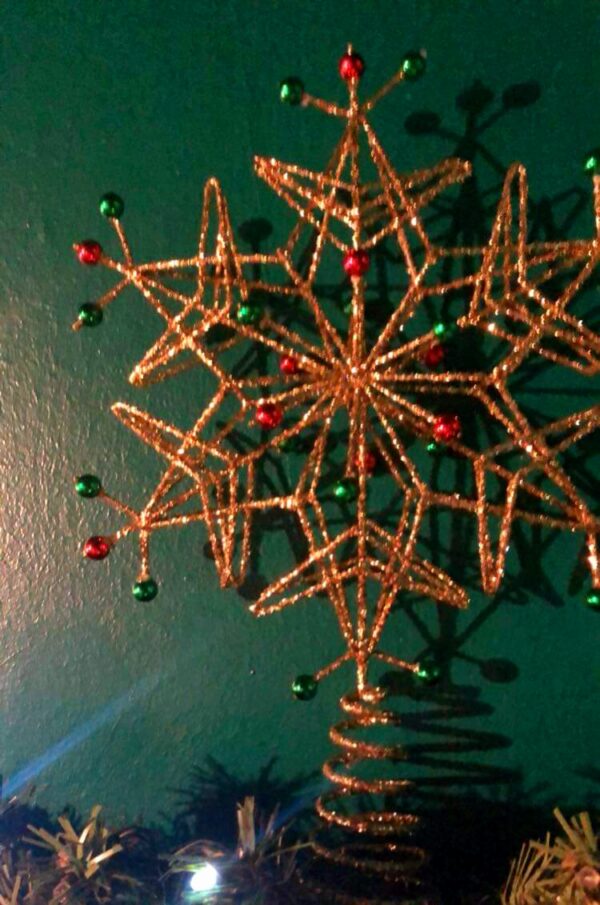 A wire frame Christmas tree topper in the shape of a six ointed snowflake. the wire frame is covered completly in gold glitter and has red and green bead on its tips and sround the centre. The snowflake sits on a conical spring for mounting on the tree.