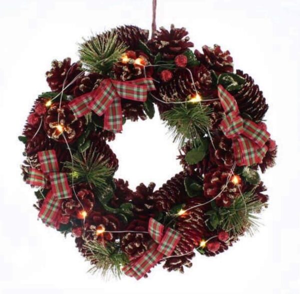 A round Christmas Wreath decorated with natural pine cones dyed red with iridescent glitter frosting, Faux pine tree sprigs and red, white and green tartan ribbon bows. Warm white LED lights are interwoven through the wreath. It hangs from a red ribbon.