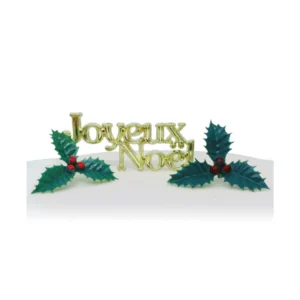 A gold plastic Christmas cake decorating motto that says Joyeux Noel that is flanked either side by two green plastic holly sprigs with red berries.