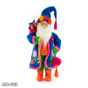 Peruvian Inspired Standing Santa Claus figurine. Santa is wearing a long blue velour coat that has colourful flowers appliqued on the sides and a pink velour trim. Beneath his coat he has an orange velour jumper and matching boots that contrast with his bright green trousers. He has a long white fluffy beard and cheerful face. On his head he wears a blue velour Santa hat with a pink tassel at the tip and a crown of colourful felt roses in red, yellow, blue and orange with green leaves. Over his right shoulder he carries a pink velour gift sack with an orange and blue wrapped present inside. In his left hand he holds a pink felt rose with an orange tassel.