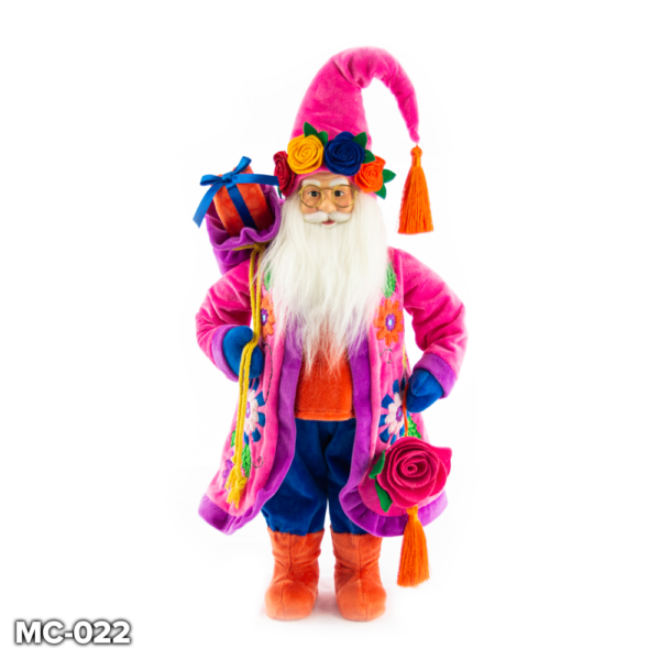 Peruvian Inspired Standing Santa Claus figurine. Santa is wearing a long pink velour coat that has colourful flowers appliqued on the sides and a purple velour trim. Beneath his coat he has an orange velour jumper and matching boots that contrast with his bright blue trousers. He has a long white fluffy beard and cheerful face. On his head he wears a pink velour Santa hat with an orange tassel at the tip and a crown of colourful felt roses in red, yellow, blue and orange with green leaves. Over his right shoulder he carries a purple velour gift sack with an orange and blue wrapped present inside. In his left hand he holds a pink felt rose with an orange tassel.