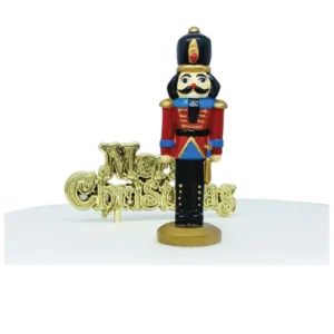 A polyresin Christmas Nutcracker cake decoration. This Nutcracker is dressed as the traditonal soldier with his red coat and black hat and trousers. He is next to a gold plastic Merry Christmas motto cake decoration.