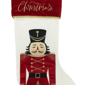 A white velour Chrsitmas stocking with an appliqued Nutcracker solider on the main body of the stocking. The Nutcracker has navy trousers and a red coat. His coat is decorated with gold embroidered eppaulettes, buttons, trim and stripes on his cuffs. his face is a peach colour with a red nose and brown eyebrows and moustache. He has black hair and is wearing a black hat with gold embroidered deatail on the from. The cuff of the stocking is red velour with gold braiding and has text embroidered across it in gold script that reads 'MERRY Christmas'. It also has a red velour loop attached to the cuff for hanging.