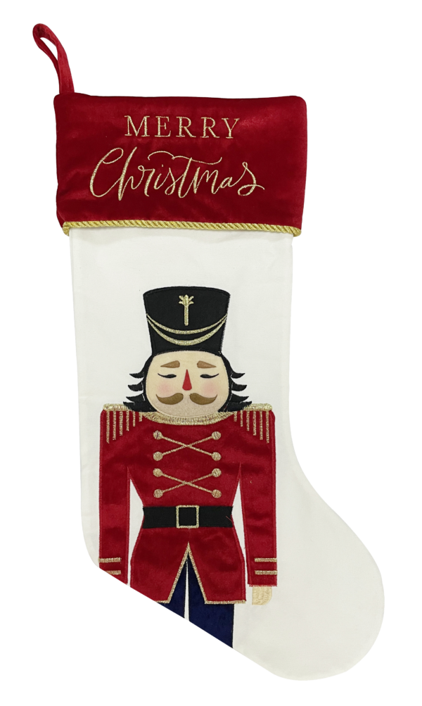 A white velour Chrsitmas stocking with an appliqued Nutcracker solider on the main body of the stocking. The Nutcracker has navy trousers and a red coat. His coat is decorated with gold embroidered eppaulettes, buttons, trim and stripes on his cuffs. his face is a peach colour with a red nose and brown eyebrows and moustache. He has black hair and is wearing a black hat with gold embroidered deatail on the from. The cuff of the stocking is red velour with gold braiding and has text embroidered across it in gold script that reads 'MERRY Christmas'. It also has a red velour loop attached to the cuff for hanging.