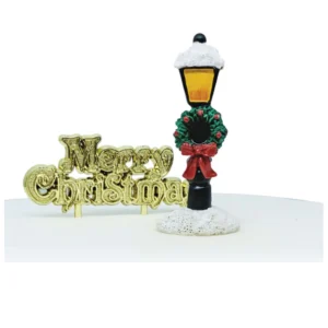 A polyresin Victorian Lamppost cake decoration. This snow topped Victorian style black lamppost has yellow windows depicting the light and is decorated with a green and red Christmas wreath with a red bow. It stands in a mound of snow and next to a gold plastic Merry Christmas cake decoration motto.