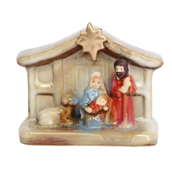 A ceramic Nativity scene depicting the manger in soft shades of cream and brown with a gold star at the top. Beneath the star Mary sits, dressed in blue robes holding the baby Jesus, who is swaddled in red cloth. On her right stands Joseph wearing long red robes. On Mary's left is a lamb looking at the baby Jesus.