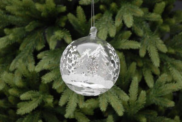 A clear glass Christmas tree bauble decorated with a winter forest scene in white and silver glitter. Hanging from a silver thread.