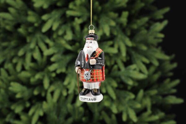 A traditional blown glass Christmas tree ornament depicting Scottish Santa in full Highland Regalia including a red tartan kilt with silver glitter sporran, black dress jacket with red tartan fly plaid over his shoulder and black Balmoral bonnet on his head. He is holding a brown pipe in his left hand and a brown walking stick in his right. He is standing on a white plinth that has the word 'SCOTLAND' across it in black text.