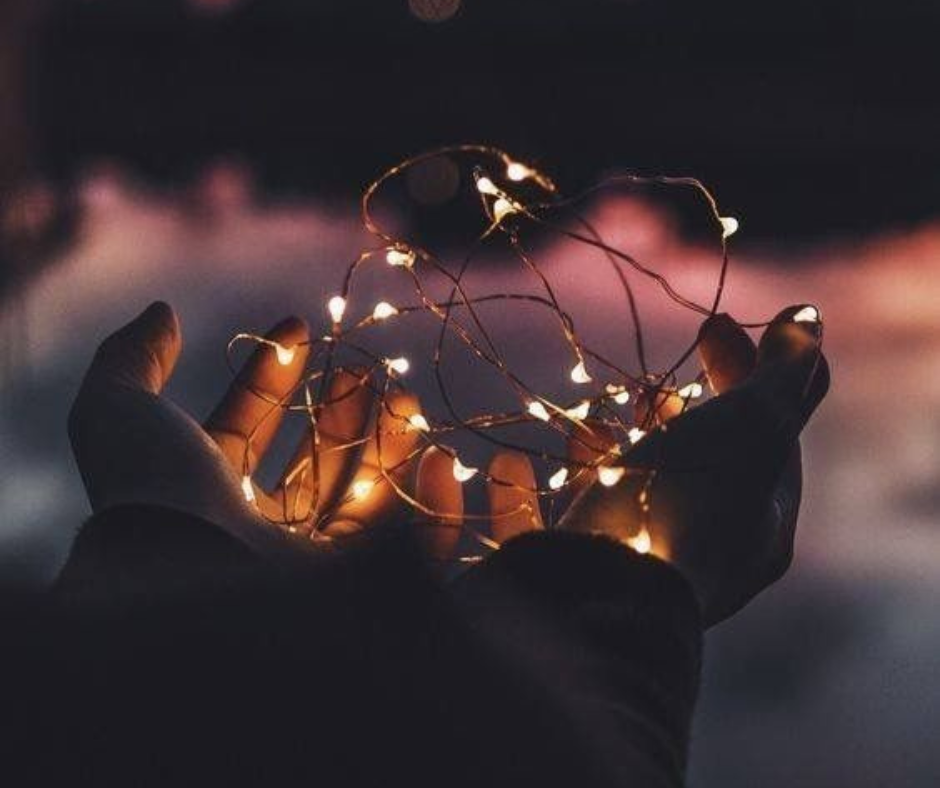 A pair of hands holding a string of lit fairy lights against a dark background