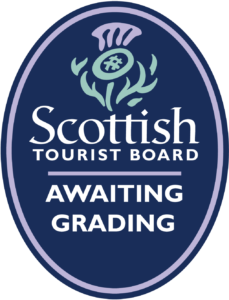 A navy oval with a pruple and green stylised Thistle at the top and white text beneath that reads 'Awaiting Grading Logo from the Scottish Tourist Board' seprated with a purple line and surrounded by a purple oval.