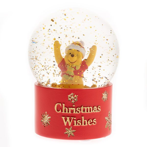 A Disney's Winnie The Pooh Snowglobe. The base of this snowglobe is red and decorated with gold snowflakes and gold text that reads 'Christmas Wishes'. Inside the globe sits a cheerful Winnie the Pooh figurine wearing a Santa hat and scarf with his arms up in the air. The globe has gold glitter inside instead of snow.