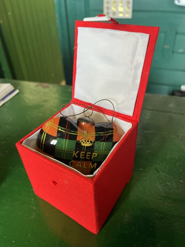 A Hand Painted Glass Heart shaped Christmas Bauble painted to look like tartan with Gift Box on a table.