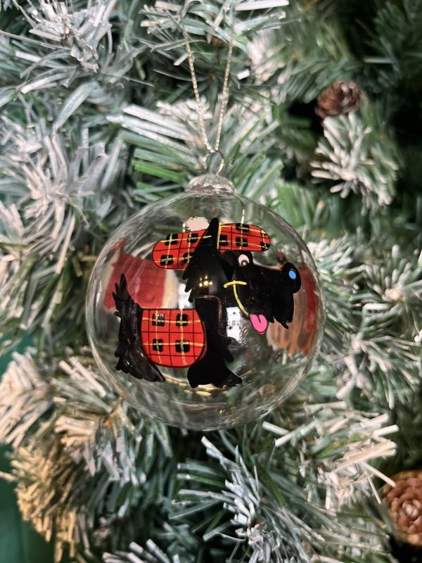 A clear glass Christmas tree bauble with a hand painted image of a black Scottie dog wearing a red Tartan coat and matching Tam O Shanter hat hanging on a Christmas tree,