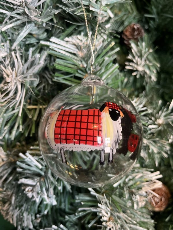 A clear glass Christmas tree bauble with a hand painted image of a white sheep wearing a red Tartan coat and matching Tam O Shanter hat hanging on a Christmas tree.