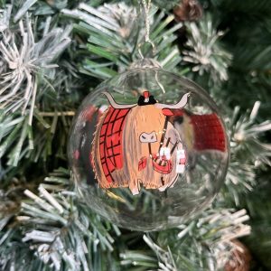 A clear glass Christmas tree bauble with a hand painted image of a Highland Cow wearing a red Tartan coat and matching Tam O Shanter hat playing the bagpipes hanging on a Christmas tree.