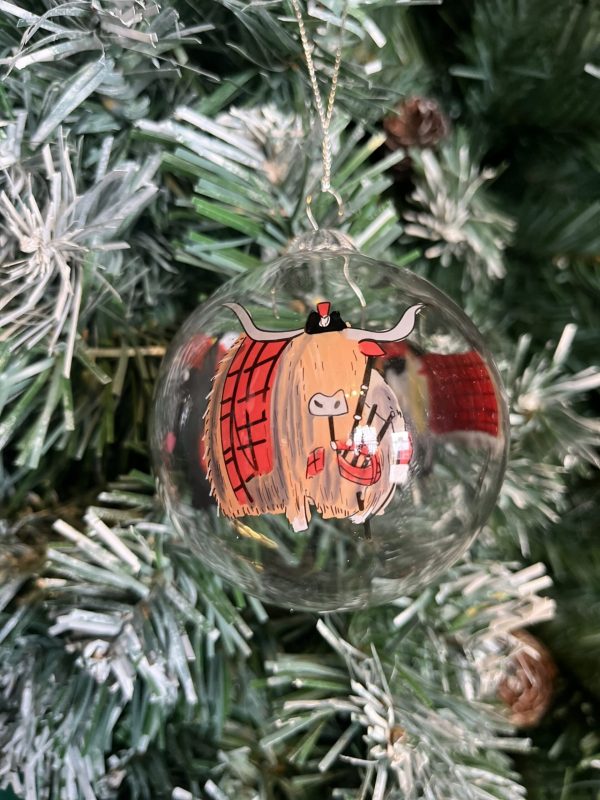 A clear glass Christmas tree bauble with a hand painted image of a Highland Cow wearing a red Tartan coat and matching Tam O Shanter hat playing the bagpipes hanging on a Christmas tree.