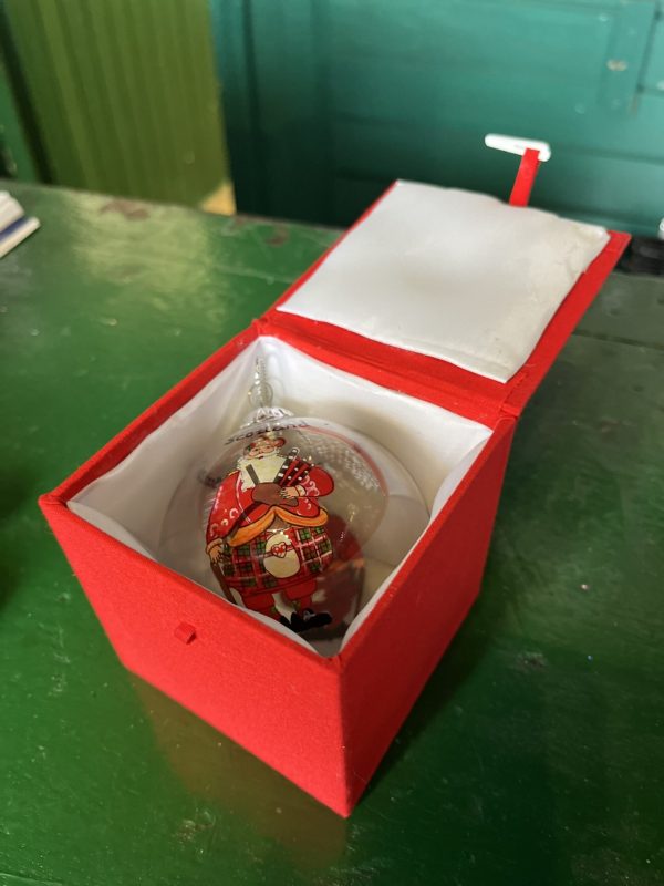 A Hand Painted Clear Glass Christmas Bauble painted with a Scottish Santa inside a red, padded Gift Box on a table.