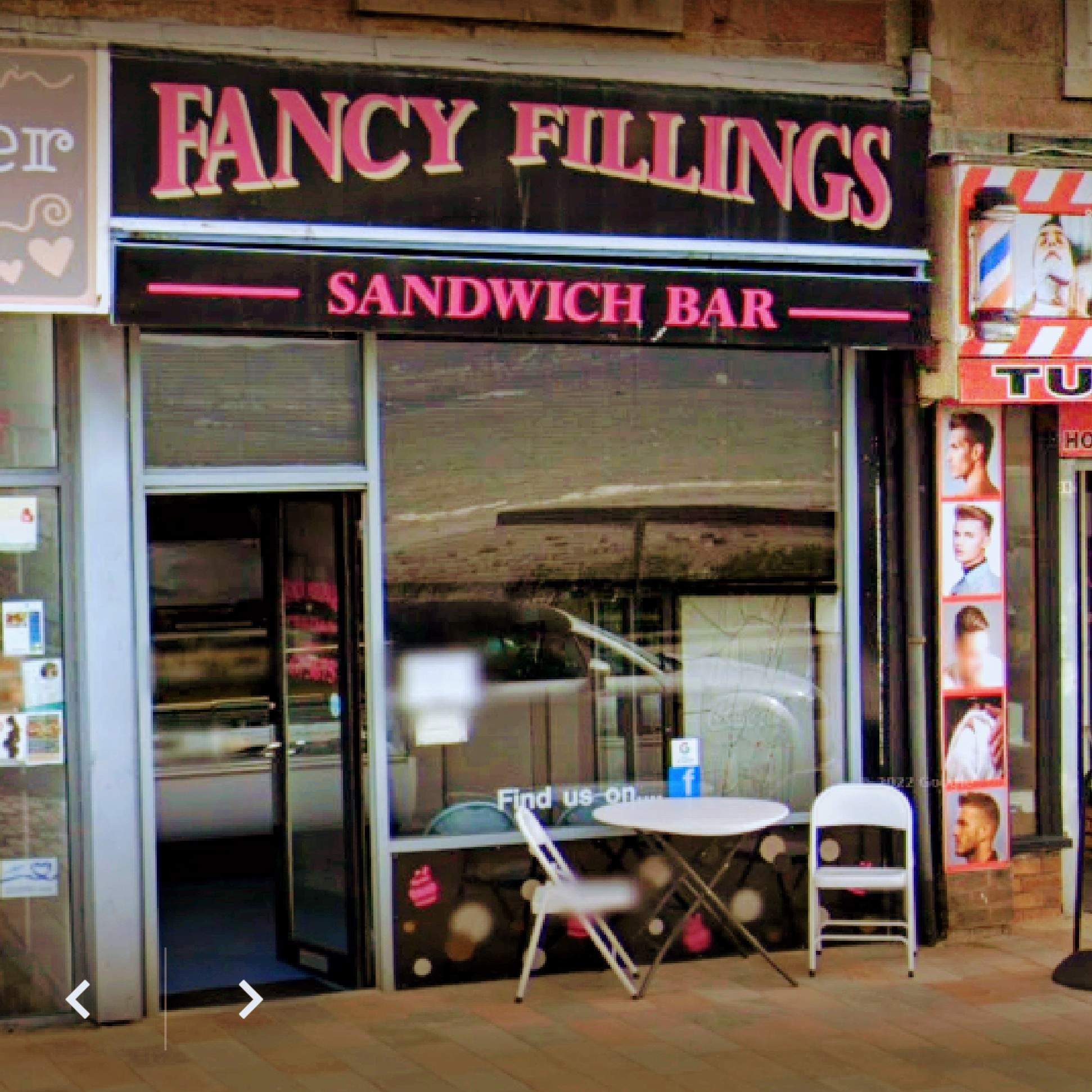 A storefront of "Fancy Fillings Sandwich Bar" with a glass door, window sign, and two white chairs with a small table outside. A barber shop is visible to the right.