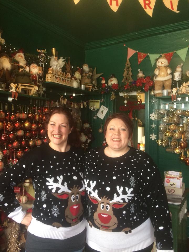 Two smiling people wearing matching reindeer sweaters stand in a festive shop filled with Christmas decorations.