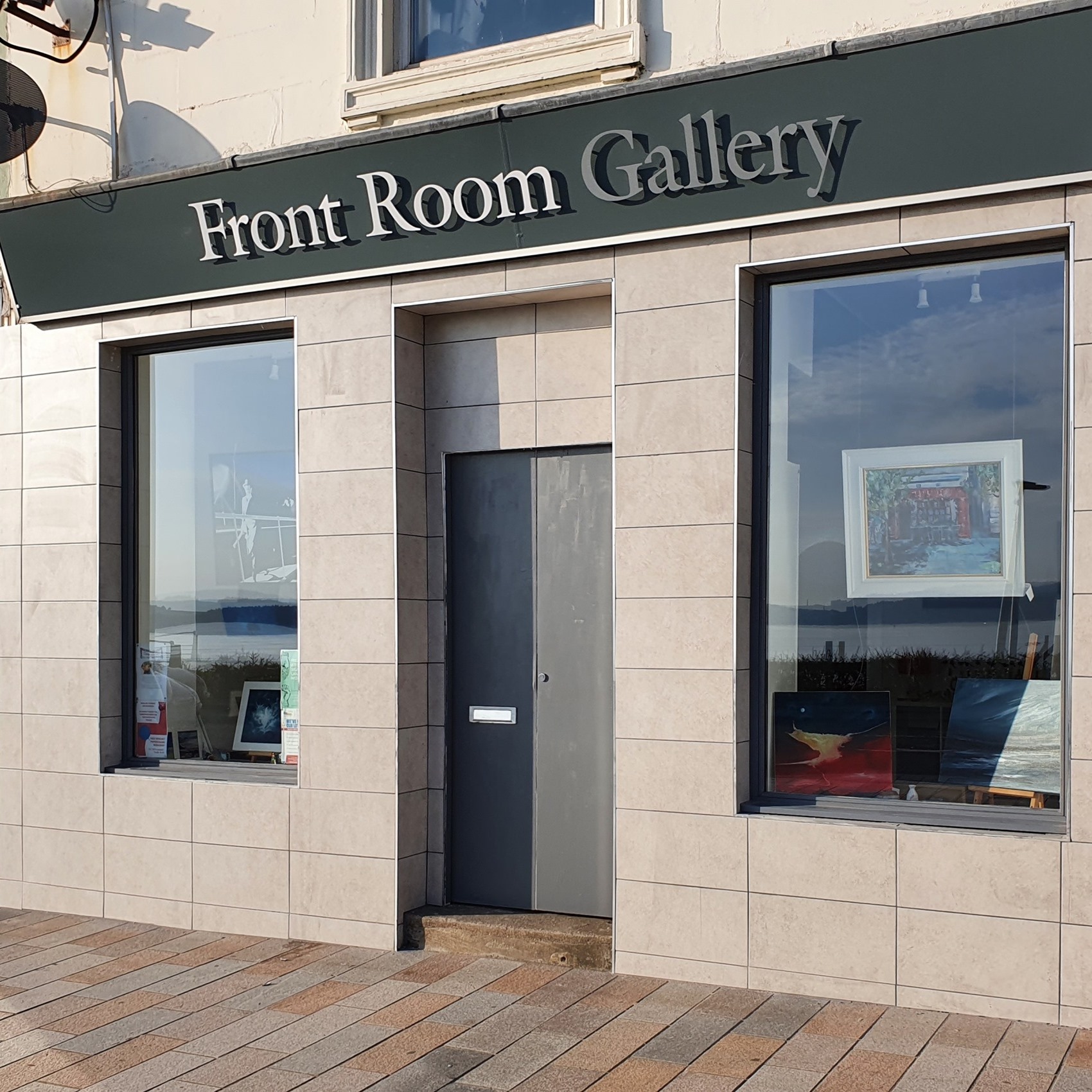 Front Room Gallery storefront with a green sign, two large display windows showcasing various artworks, and a gray door in the center on a tiled facade.